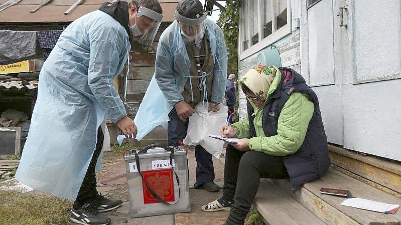 Zwei Wahlhelfer stehen neben einer Frau, die auf den Stufen vor einem Haus sitzt und Dokumente ausfüllt. Foto: Evgeniy Sofiychuk/AP/dpa