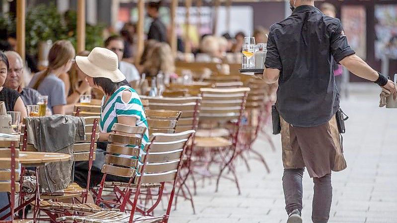 Eine Servicekraft läuft im Außenbereich eines Restaurants an Gästen vorbei. Foto: Christoph Schmidt/dpa