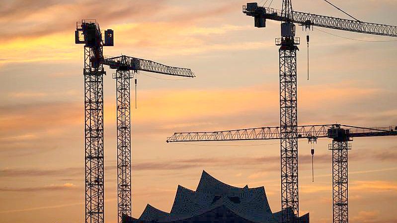 Die Kräne verschiedener Baustellen in der Hamburger Hafencity zeichnen sich im Sonnenuntergang vor der Elbphilharmonie ab. Foto: Christian Charisius/dpa