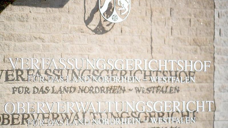 An der Außenfassade am Oberverwaltungsgericht hängt eine Hinweistafel mit dem Landeswappen von NRW. Foto: Guido Kirchner/dpa