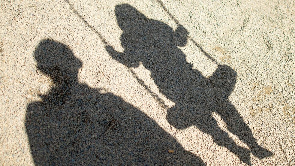 Der Schatten von einem Mann und einem schaukelnden Kind fallen auf Sand auf einem Spielplatz. Foto: DPA