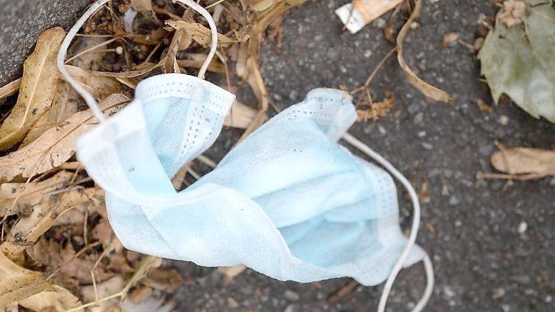 Eine Schutzmaske liegt neben Laub und einer Zigarettenkippe auf dem Asphalt. ). Foto: Marijan Murat/dpa