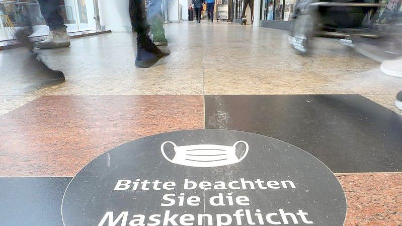 Passanten laufen durch ein Einkaufszentrum auf dessen Fußboden ein Hinweisschild zur Maskenpflicht aufgeklebt ist. Foto: Danny Gohlke/dpa