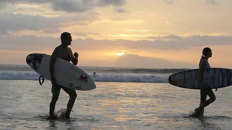 Surfer gehen an einem Strand auf Bali durch seichtes Wasser. Foto: Firdia Lisnawati/AP/dpa/Archivbild