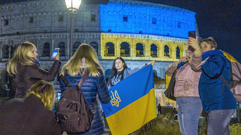 Menschen stehen vor dem Kolosseum in Rom, das in den Farben der ukrainischen Nationalflagge beleuchtet ist, um sich mit der ukrainischen Bevölkerung zu solidarisieren. Foto: DPA