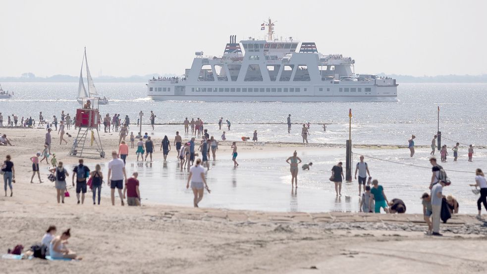 Eine Fähre der Reederei Frisia fährt im Jahr 2019, vor Beginn der Pandemie, an einem Strand vorbei, an der Urlauber und Einheimische einen Sommertag genießen. Foto: DPA
