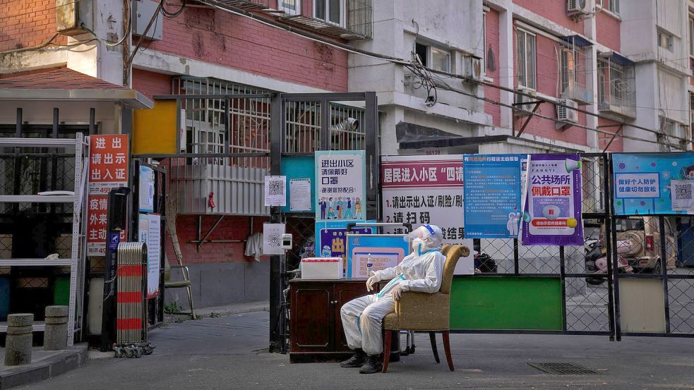 Ein Wachmann im Schutzanzug sitzt an einem Eingangstor zu einem Wohnviertel in Peking. Foto: Andy Wong/AP/dpa