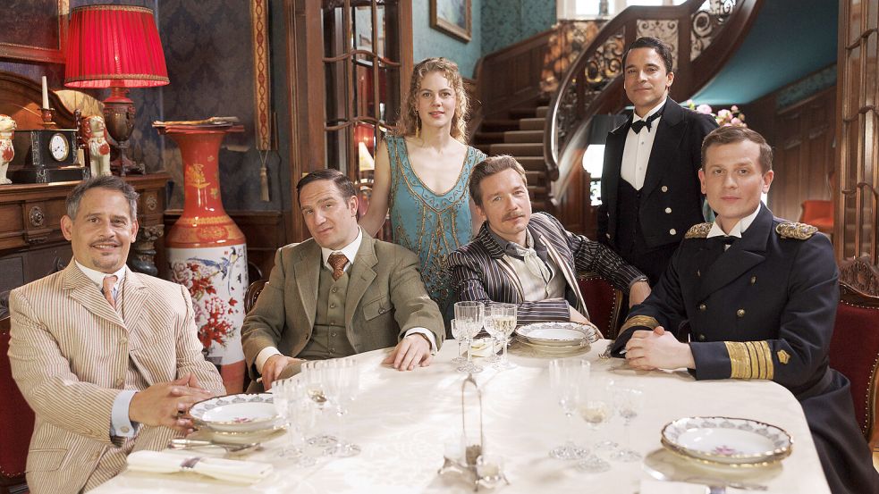 „Dinner for one“ wird zum „Dinner for five“: Am gedeckten Tisch sitzen Moritz Bleibtreu als Mr. Pommeroy, Frederick Lau als Mr. Winterbottom, Alicia von Rittberg als Miss Sophie, Jacob Matschenz als Sir Toby, Kostja Ullmann als Butler James sowie Christoph Schechinger als Admiral von Schneider. Foto: Prime Video/Frédéric Batier