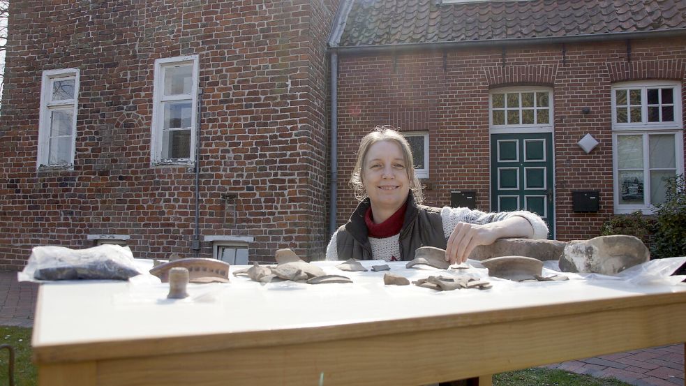Archäologin Dr. Sonja König zeigt im Frühjahr 2021 die keramischen Überreste, die bei der Ausgrabung am Gemeindehaus Engerhafe ans Licht gekommen waren. Im Hintergrund sind das damals noch nicht sanierte Pfarrhaus und das Gemeindehaus zu erkennen.