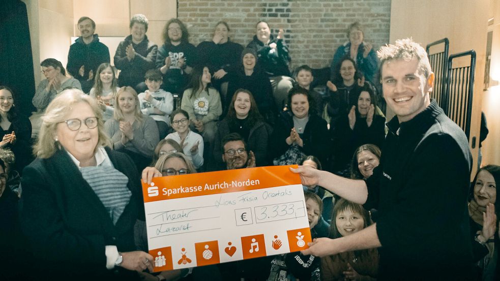 Eva Decking, Präsidentin des Auricher Lions-Clubs Frisia Orientalis, übergibt einen symbolischen Scheck an Eike Schmidt, Gründer des Theater Lazarett. Im Hintergrund applaudieren Kinder und Jugendliche vom inklusiven Jugendtheater Familie Gassenhauer. Foto: Privat